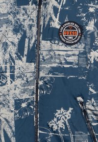 Tmavě modrá bunda na zip z polyesteru s bílým květinovým a palmovým potiskem. Má našitou nášivku s logem a boční kapsu na zip.