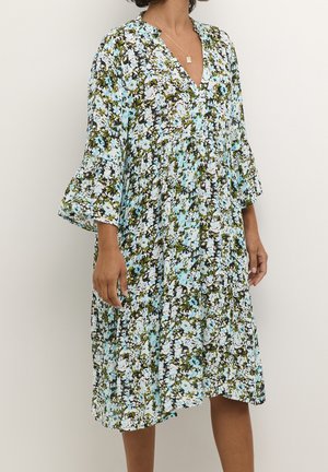 Frau trägt ein knielanges Kleid mit blauen, weißen und grünen Blumenmustern und 3/4 Glockenärmeln, stehend vor einfachem Hintergrund.