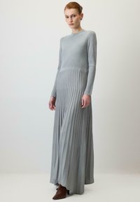 Kvinde iført en lang, lysegrå ribstrikkjole med lange ærmer og brune spidse ankelstøvler, der står op ad en ensfarvet baggrund.