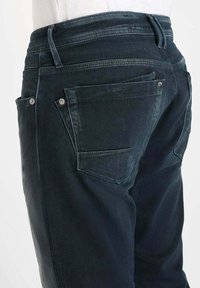 Jean en denim foncé avec une texture légèrement dégradée, design classique à cinq poches, accents en métal, et taille haute avec des coutures décoratives.
