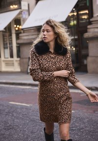 Robe imprimé léopard marron avec col en fausse fourrure noire, manches longues, boutonnée à l'avant et coupe jusqu'au genou. Portée avec des bottes noires.