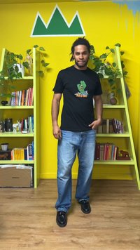 Musta t-paita, jossa vihreä graafinen kuva kädestä, yhdistetty vaaleansiniin farkkuihin ja mustiin kenkiin. Tausta on keltainen, jossa vihreitä yksityiskohtia.