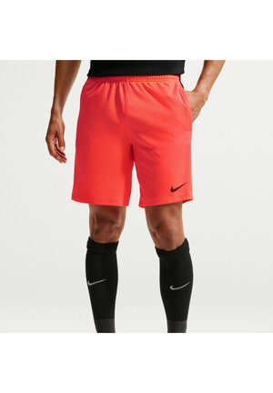 Homme portant un short de sport rouge vif Nike et des chaussettes noires montantes Nike, debout avec une main dans la poche.