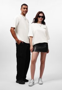Weiße übergroße T-Shirts, schwarze Cargo-Hosen und einen schwarzen Leder-Minirock. Zubehör umfasst Sonnenbrillen und Halsketten. Sneakers runden den Look ab.