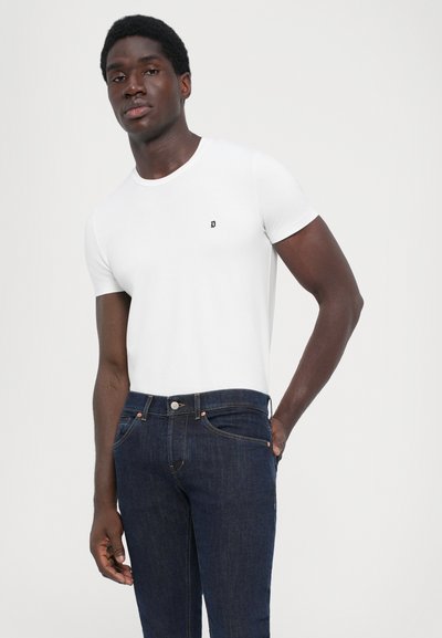 Camiseta blanca de manga corta con un pequeño logotipo negro en el pecho, combinada con unos pantalones vaqueros de mezclilla azul oscuro que presentan un diseño tradicional de cinco bolsillos.