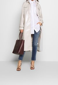 Henkilö, jolla on päällään beige pitkä takki, valkoinen paita, siniset farkut ja korkokengät, pitää kädessään suurta tummanruskeaa olkalaukkua.