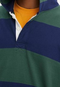 Primo piano di abbigliamento a strati: felpa con cappuccio a righe verdi e blu navy sopra una camicia bianca con bottoni e una maglietta gialla senape.