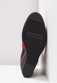 Podeszwa buta Calvin Klein Jeans, wykonana z czarnej gumy z poziomymi paskami i logo, obok czerwono-czarnego materiałowego segmentu cholewki.