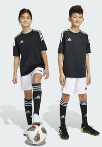 Schwarz-weiße Sportshirts mit weißen Streifen, weiße Shorts, schwarze kniefreie Socken mit grauen Streifen und Fußballschuhe. Ein Fußball liegt zwischen ihnen.
