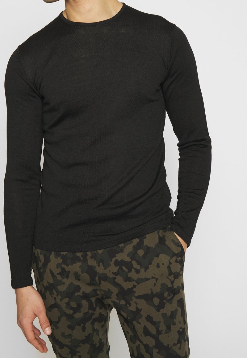 Homme portant une chemise noire ajustée à manches longues avec un pantalon camouflage vert foncé et noir, une main dans la poche, sur un fond uni.