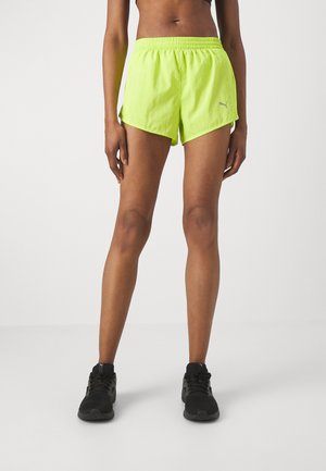 Pantalones cortos deportivos de color verde neón con un cinturón elástico, aberturas laterales y un pequeño logo reflectante en la parte inferior izquierda. Tela ligera.