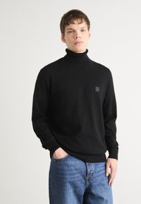 Črn pulover s polo ovratnikom, izdelan iz mehkega blaga, s rebrastim ovratnikom in manšetami. Majhen kvadraten logotip je na levem prsnem delu. Nosite ga s svetlo modrimi kavbojkami.