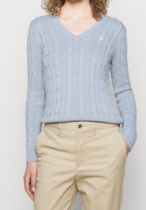 Kobieta ubrana w jasnoniebieski sweter z warkoczowym wzorem i dekoltem w serek oraz beżowe spodnie, z rękami w kieszeniach, na jednolitym tle.