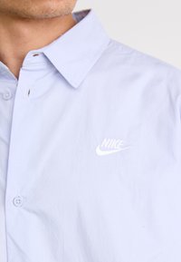 Lys blå skjorte med knappemynomd, utstyrt med en hvit Nike-logo, laget av glatt stoff med klassisk krage og subtil tekstur.