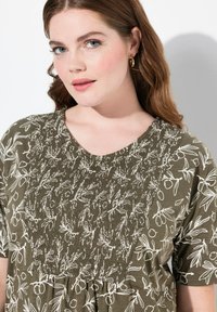 Haut vert olive avec impression florale blanche. Présente un corps smocké et des manches courtes, créant une coupe décontractée et une surface texturée.