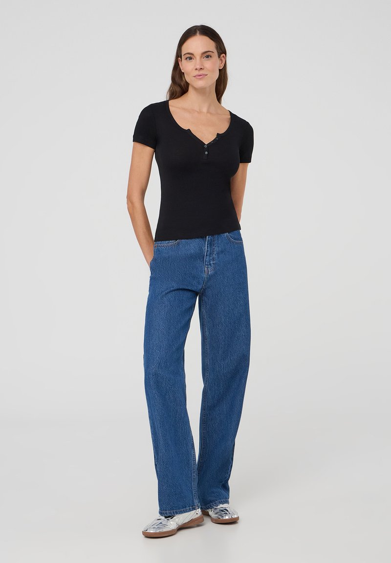 Top nero a maniche corte con scollo a V e dettagli in bottone, abbinato a jeans azzurri a vita alta e gamba larga, e scarpe metalliche.