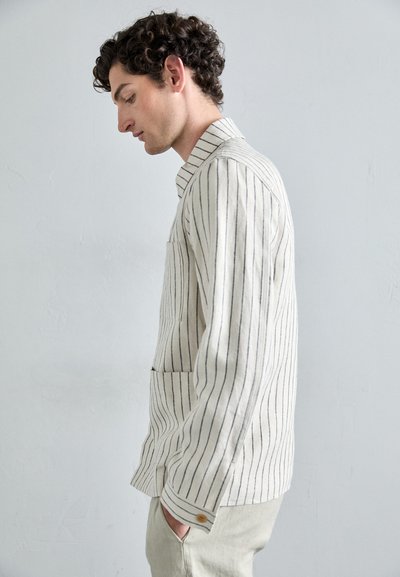 Chemise rayée en beige clair et bleu marine, en tissu texturé, avec un col pointu et une poche frontale. Modèle de profil.