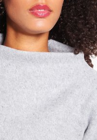 Tissu polaire gris avec un col plié, présentant une texture douce. Les lèvres sont légèrement teintées de rose.