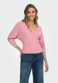 Różowy sweter z prążkowanego dzianiny z głębokim dekoltem w serek, dopasowaną talią i długimi rękawami, w połączeniu z niebieskimi jeansami. Gładka faktura, miękki materiał.