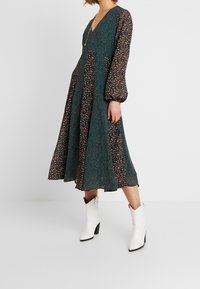 Dunkelgrünes Kleid mit Blumen- und Punktmuster, V-Ausschnitt, langen Ballonärmeln und einem fließenden Saum, kombiniert mit weißen Absatzstiefeln.