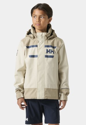 Jongen draagt een beige en bruine waterdichte jas met capuchon en marineblauwe korte broek, en kijkt opzij tegen een effen achtergrond.