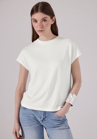 Bílý krátký rukávový top s uvolněným střihem a kulatým výstřihem, zkombinovaný se světle modrými džíny a stříbrným náramkem. Hladká textura.