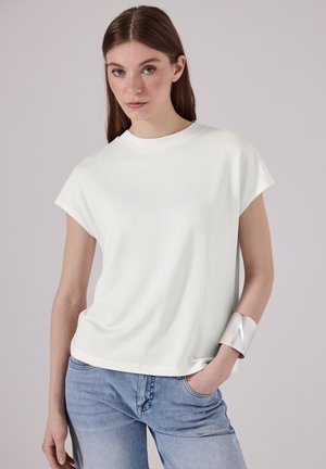 Haut blanc à manches courtes avec une coupe décontractée et un col rond, associé à un jean bleu clair et un bracelet en argent. Texture lisse.