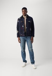 Σκούρα μπλε τζιν μπουφάν με λουλουδάτες κεντήσεις, μαύρο γιακά, μπροστινές τσέπες; συνδυασμένο με ανοιχτό μπλε τζιν και άσπρα αθλητικά παπούτσια.