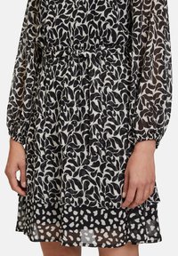 Schwarz-weiß gemustertes Kleid mit floralen Designs, durchsichtigen langen Ärmeln, gerafften Bündchen und einem geschichteten, rüschierten Saum. Das Material wirkt leicht.