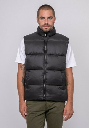 Homme aux cheveux courts portant un gilet matelassé noir sur un t-shirt blanc et un pantalon vert olive, debout devant un fond gris uni.