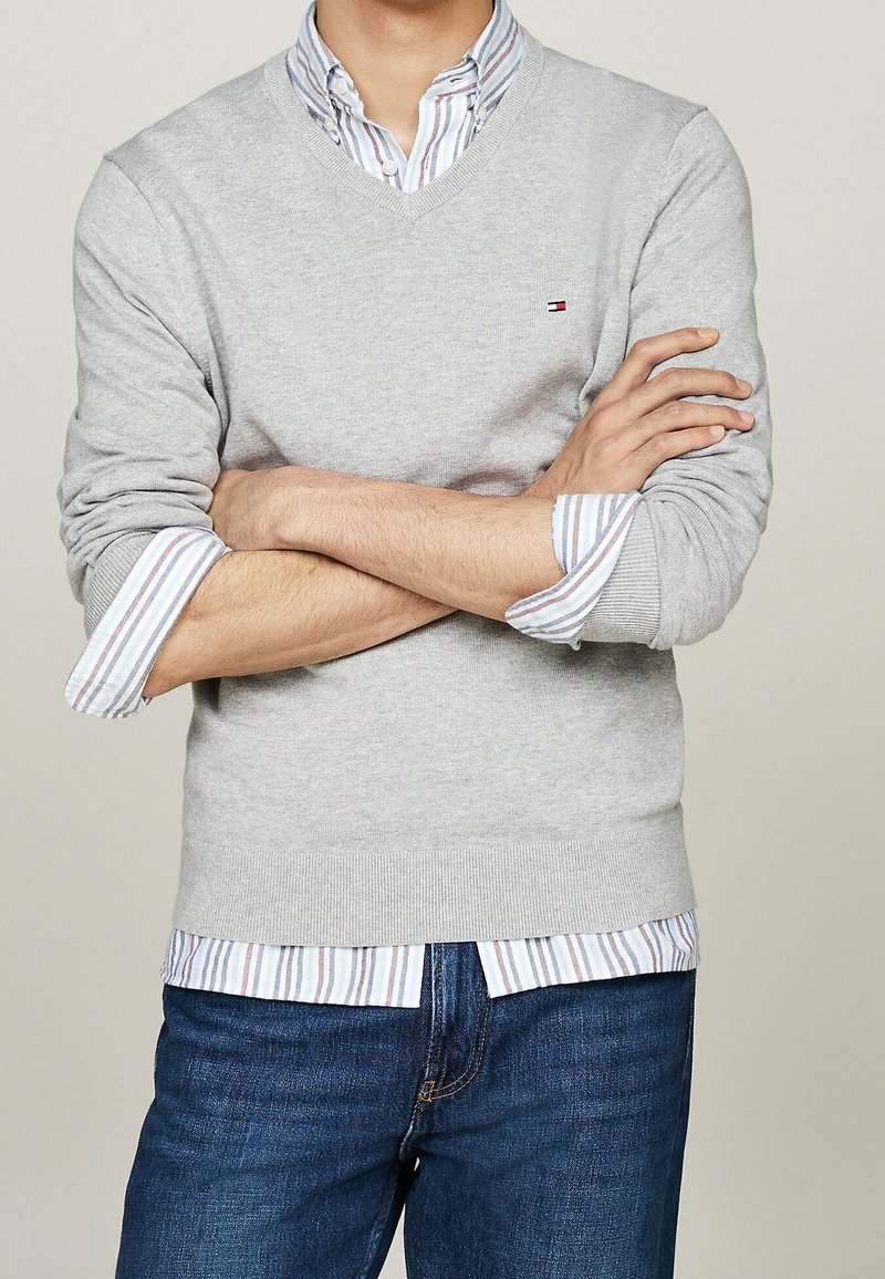 Homme les bras croisés portant un pull gris clair sur une chemise à rayures boutonnée et un jean bleu, sur fond uni.
