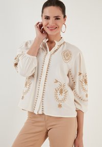 Valkoinen pitkähiha blouse, jossa on monimutkaista beigeä brodeerausta, röyhelökaulus ja napit edessä, sekä puffihihat.
