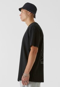 Black cotton t-shirt with a relaxed fit, short sleeves, and graphic detail on the back. Worn with a navy bucket hat, showing a side profile.