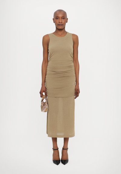 Rochie bodycon fără mâneci, de culoare khaki, cu detalii de ruflare, design lungime până la glezne și o secțiune inferioară transparentă, asortată cu o geantă mică cu model și tocuri negre.