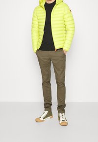 Neon žlutá péřová bunda s vodorovným prošíváním, spojená s černým tričkem a olivově zelenými slim-fit kalhotami. Bílé a zelené tenisky.