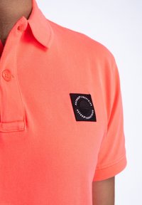 Korálová polo košile s límečkem, se čtvercovou černou nášivkou s kulatým designem a textem. Textura vypadá hladce s jemným tkaním.