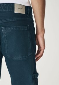 Denim jeans in donker teal. Kenmerken zijn een achterzak met dubbele stiknaad en een label met de tekst "DENIM SUPPLY." Soepele textuur.