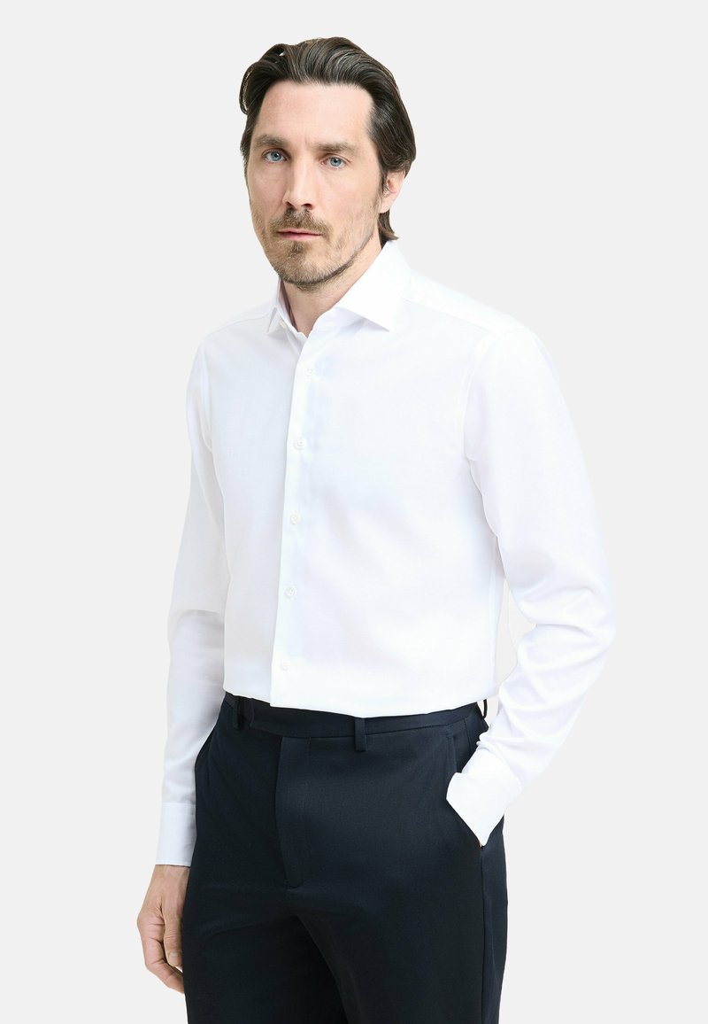 Man met donker haar en baard, gekleed in een witte blouse en zwarte broek, staande met één hand in de zak tegen een eenvoudige achtergrond.