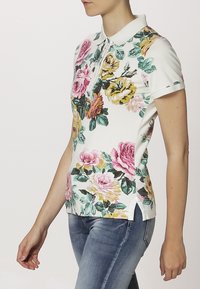 Polo à manches courtes en blanc avec un motif floral comprenant des roses roses et jaunes, ainsi que des feuilles vertes. Tissu doux et texturé avec des fentes sur les côtés.