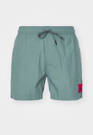 Zöld úszónadrág rugalmas derékpánttal és húzózsinórral. A bal oldalon egy piros logópatccsal rendelkezik, könnyű anyagból készült.