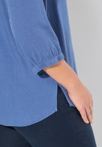 Blusa leggera blu con maniche a tre quarti, polsini arricciati e fessure laterali, abbinata a pantaloni blu scuro. Tessuto testurizzato.
