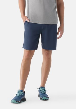 Person, die marineblaue Shorts, graues T-Shirt und blaue Sportschuhe trägt, steht mit einer Hand in der Tasche vor weißem Hintergrund.