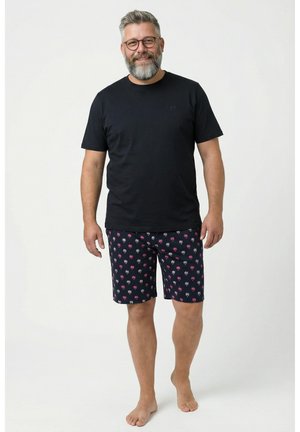 Brodaty mężczyzna w okularach, ubrany w czarny t-shirt i granatowe szorty w różowo-białe wzory palm, stoi boso na jednolitym tle.