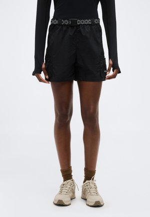 Person trägt schwarze Cargo-Shorts mit gemustertem Gürtel, schwarzes Langarmshirt, braune Socken und beige Turnschuhe und steht vor weißem Hintergrund.