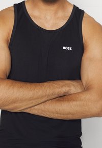 Černý tílko vyrobené ze hladkého materiálu, s kulatým výstřihem a malým bílým logem "BOSS" na hrudi. Bez viditelných vzorů.
