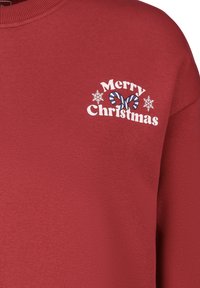 Rotes Sweatshirt mit weißem Text "Frohe Weihnachten" und einem Schmetterlingsdesign, akzentuiert durch weiße Schneeflocken und Zuckerstangenelemente.