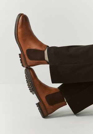 Braune Leder-Chelsea-Stiefel mit schwarzen elastischen seitlichen Einsätzen, getragen mit schwarzen Hosen und hellbeigen Socken, fotografiert vor einem einfarbigen Hintergrund.