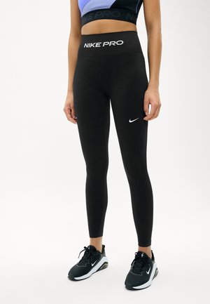 Personne portant un legging taille haute Nike Pro noir et des baskets Nike noires, debout devant un fond clair uni.