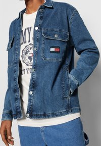 Giacca di denim blu medio con chiusura a bottoni, doppie tasche sul petto e etichetta con logo che mostra rosso, bianco e verde. Tessuto morbido e texturizzato.