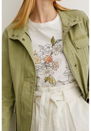 Kobieta ubrana w jasnozieloną kurtkę na białej koszulce z kwiatowym nadrukiem na twarzy oraz białe spodnie z wysokim stanem i zawiązanym paskiem.